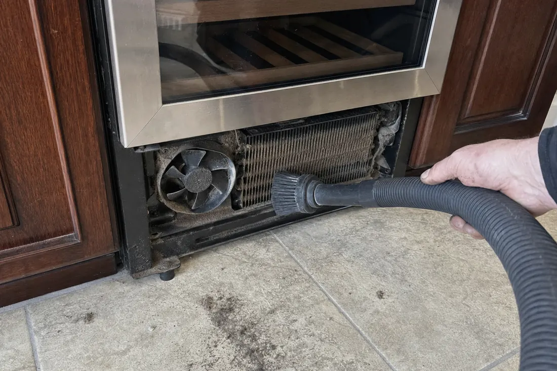 A brush and vacuum hose on the dust-loaded condenser coil behind a removed grille, the cabinet doors either side in frame - Summer heat load, and the condensers that give up