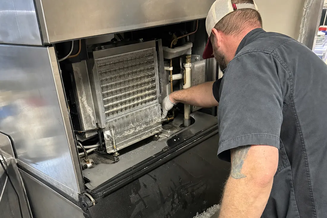 Technician at a stainless ice machine with the top panel off, checking the water line and the evaporator plate, the water line in frame - Ice machine and ice maker repair