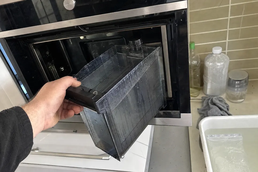 Hands lifting the water reservoir out of a built-in steam oven, the descaling supplies in frame - Miele appliance repair