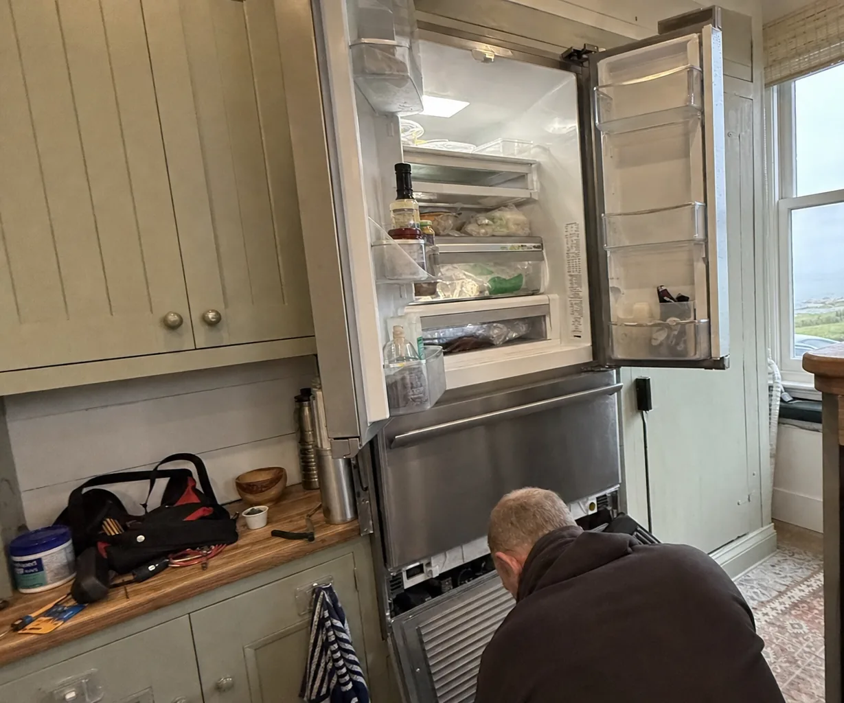 Technician kneeling at the open lower grille of a built-in French-door refrigerator with both doors open, sage-green cabinets and a tool bag on the counter - Built-in refrigerator design and service access