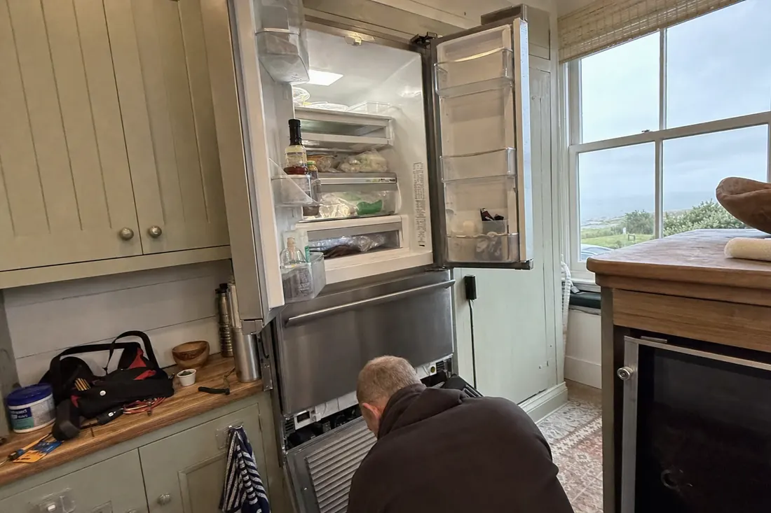 An open built-in refrigerator in a coastal kitchen, technician at the lower grille, the lower grille in frame - Six badges. One weather system.