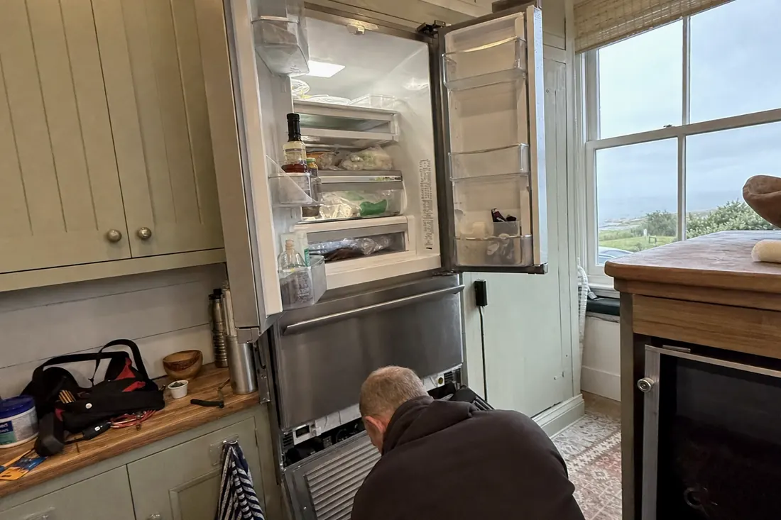 An open built-in refrigerator in a coastal kitchen, technician at the lower grille, the painted timber cabinets in frame - Appliance repair in Orange Park and Fleming Island
